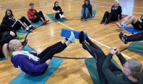 Eine Gruppe von angehenden Co-Trainer*innen sitz in einem Sitzkreis zusammen. Zwei Teilnehmende halten gemeinsam ein blaues Pad mit ihren Füßen in die Luft.