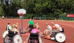 Kinder stehen in Rollstühlen sitzend im Kreis um einen Basketballkorb.