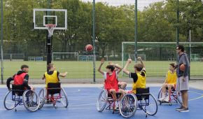 Sechs Jungs spielen Rollstuhlbasketball. Der Ball fliegt durch die Luft. Der Trainer steht beobachtend daneben und hält einen weiteren Ball in der Hand