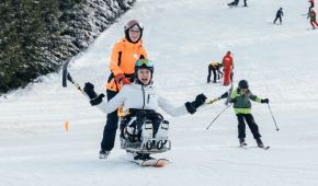 Zwei Skifahrer*innen fahren den Berg herunter, eine Person sitzt im Bi-Skigerät