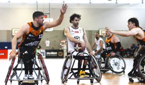 Mehrere Rollstuhlbasketballspieler duellieren sich um den Ball. Der Spieler in der Mitte hält ihn fest und wird von zwei Gegnern angegriffen