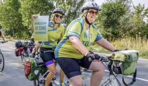 Zwei Frauen fahren auf einem Tandem. Beide tragen ein Shirt der Mut-Tour, eine Frau hält ein Schild mit der Aufschrift: Depression ist einer Erkrankung