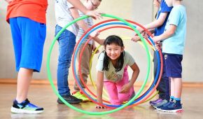 Mehrere Kinder spielen in einer Turnhalle. Fünf Kinder halten hintereinander mehrere Reifen fest, ein Mädchen klettert durch diese durch.