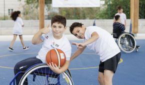 Ein Junge sitzt im Rollstuhl und hat einen Basketball in der Hand, bei der anderen Hand zeigt der Daumen nach oben. Dahinter ist ein zweiter Junge, der das Peace-Zeichen zeigt. Im Hintergrund spielen weitere Kinder Basketball, teilweise im Rollstuhl.
