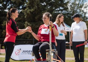Szene beim Jugend-Länder-Cup: Ein Mädchen, reicht einem anderen Mädchen, das auf einem Wurfstuhl sitzt, die Kugel.