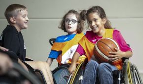 Spielszene beim Rollstuhlbasketball mit drei Kindern in der Sporthalle