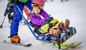 Ein junges Mädchen sitzt in einem Para Ski Schlitten und lacht in die Kamera