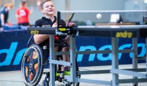 Ein Junge sitzt in seinem Rollstuhl vor der Tischtennisplatte und hält seinen Schläger bereit